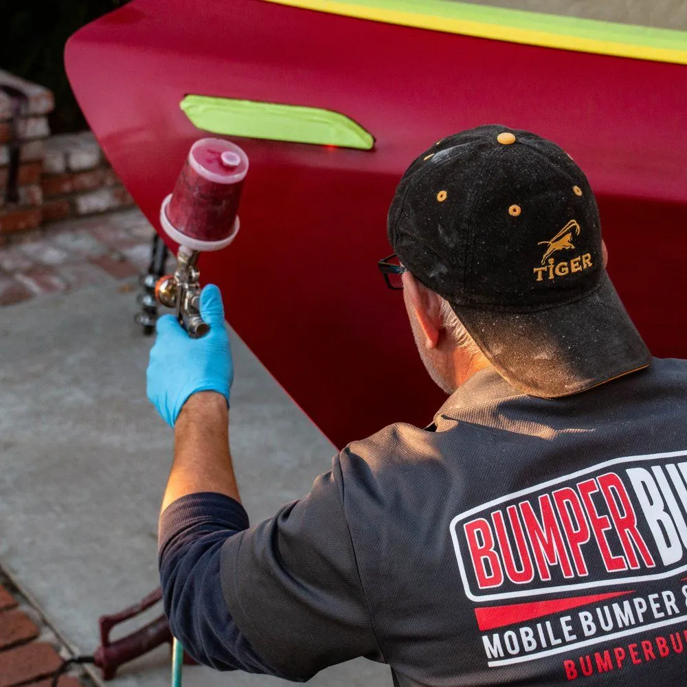 Worker spray-painting a red bumper car with a purple spray gun, wearing blue gloves and a black cap.
