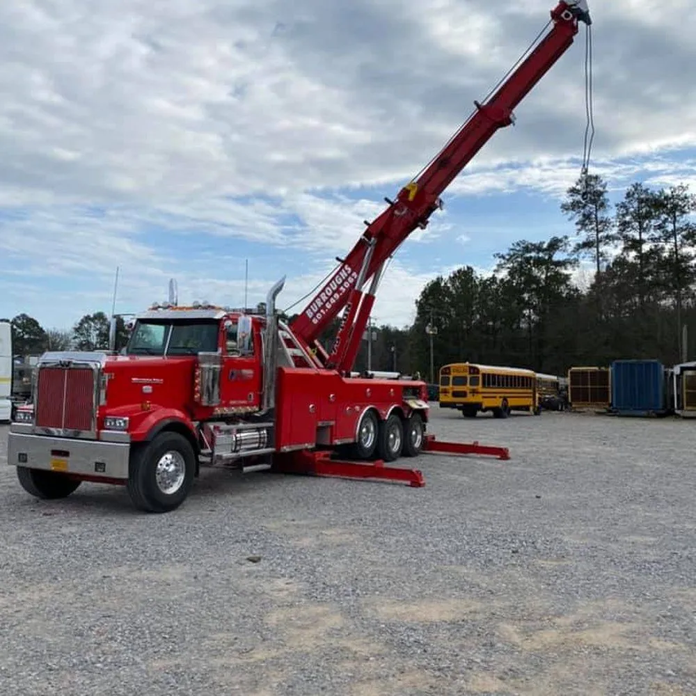 Red tow truck with raised boom in a gravel lot, with school buses and a trailer in the background