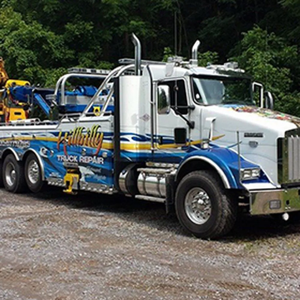 Blue-and-white heavy-duty tow truck on a gravel lot, labeled "truck repair," with a forested background