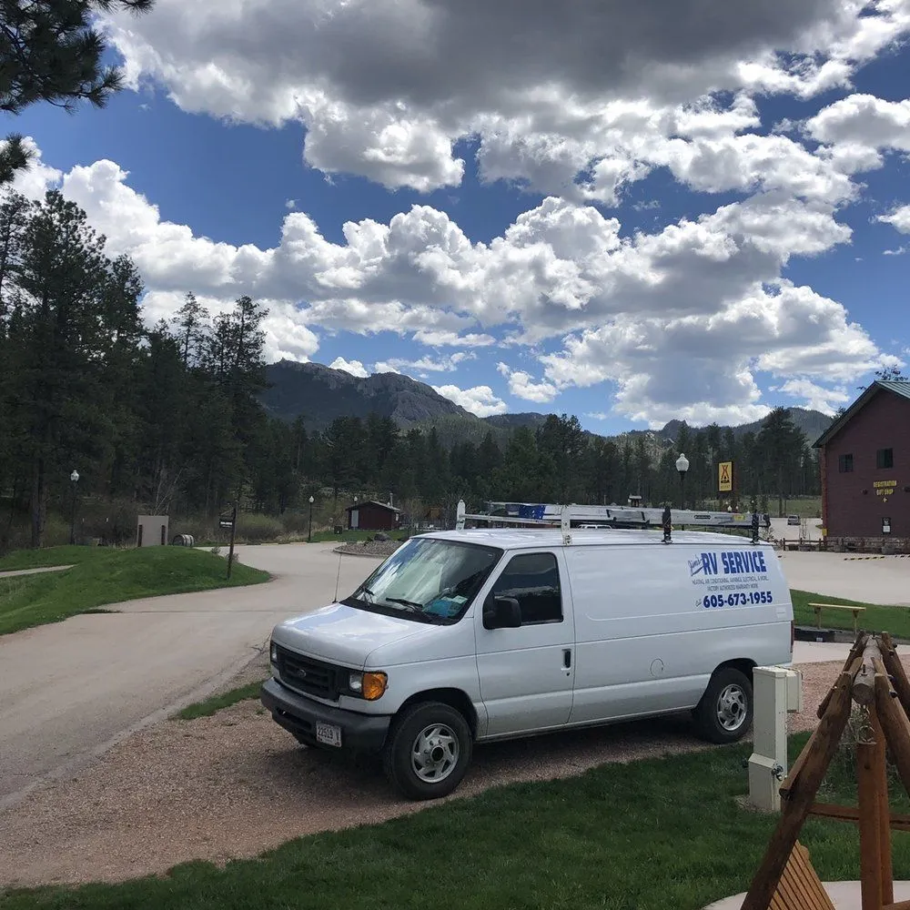 White utility van parked on a mountain road under a cloudy sky, with trees and cabins nearby