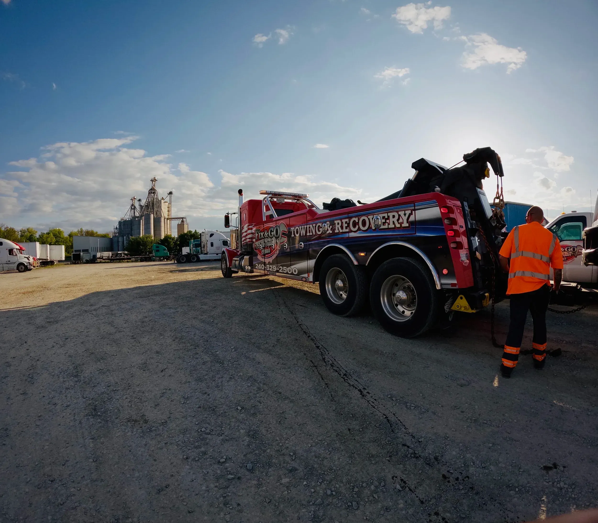 Tow truck driver in orange safety gear beside a red tow truck on a gravel lot at dusk