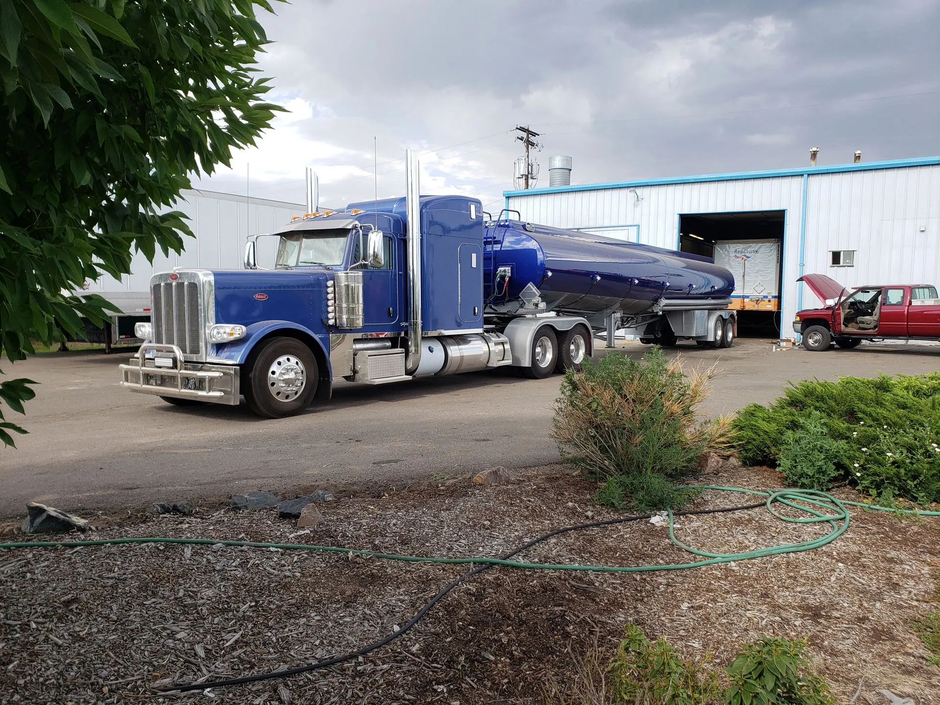 Blue semi-truck hauling a tanker trailer parked outside a warehouse on a gravel lot